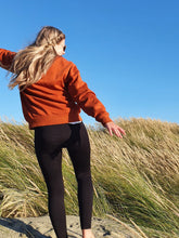 Load image into Gallery viewer, Here is Zoey sporting our Signature Rust Crew on the couch. This is made of Organic Cotton and Recycled polyester. Zoey wears a Medium so that she can fit a shirt or long sleeve comfortably underneath.