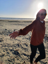 Load image into Gallery viewer, Here is Zoey sporting our Signature Rust Crew on the couch. This is made of Organic Cotton and Recycled polyester. Zoey wears a Medium so that she can fit a shirt or long sleeve comfortably underneath.